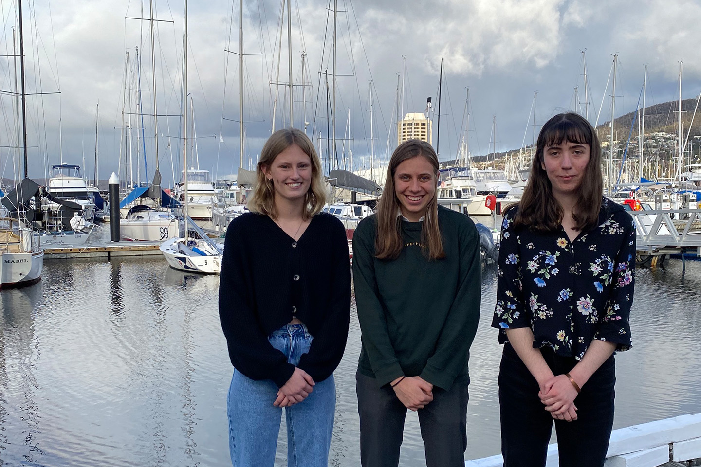 Three women standing for a photo in a marina with sail boats behind them, all smiling